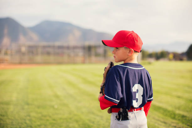Little League Baseball Boy Profile A young Little League baseball player is wearing a baseball uniform and holding his baseball glove while standing in the outfield. The boy loves playing America's pastime on warm summer days in Utah, USA. baseball uniform photos stock pictures, royalty-free photos & images