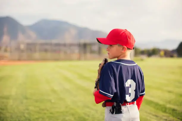 A young Little League baseball player is wearing a baseball uniform and holding his baseball glove while standing in the outfield. The boy loves playing America's pastime on warm summer days in Utah, USA.