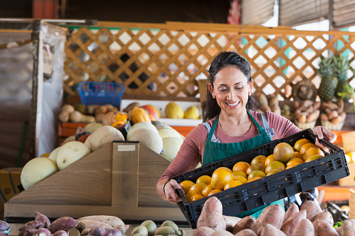 A mature Hispanic woman in her 40s working at a farmer's market produce stand. She is looking down at the crate of oranges she is holding.