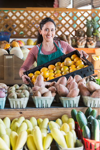 A mature Hispanic woman in her 40s working at a farmer's market produce stand. She is looking at the camera, holding a crate of oranges.