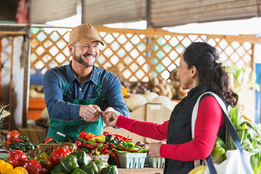 A mature man working at a farmer's market produce stand, smiling as he accepts payment from his customer, an Hispanic woman.