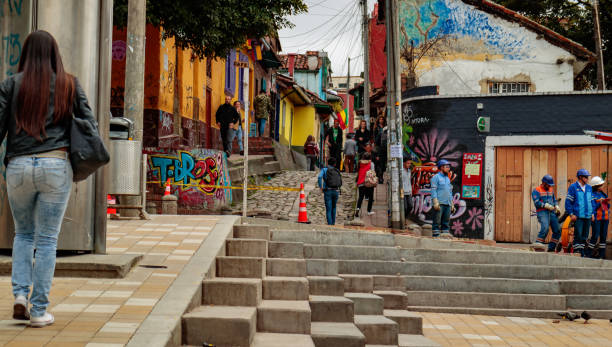 Bogota, Colombia - Looking Towards the Calle del Embudo in the Historic La Candelaria District of the Andean Capital City Bogota, Colombia - January 27, 2017: Looking towards Calle del Embudo, one of the most colorful, old streets in the historic La Candelaria District on the Andean capital city of Bogota in Colombia, in South America. The street leads to the Chorro de Quevedo, the plaza where It is believed, the Spanish Conquistador, Gonzalo Jiménez de Quesada founded the city in 1538. Many walls in this area are painted with either street art, or legends of the pre Colombian era, in the vibrant colours of Latin America. Some old colonial buildings can be seen as well. Some local Colombian people are seen going about their daily chores while some workers are engaged in laying some cables. Photo shot on a cloudy morning; horizontal format. Copy space. calle del embudo stock pictures, royalty-free photos & images