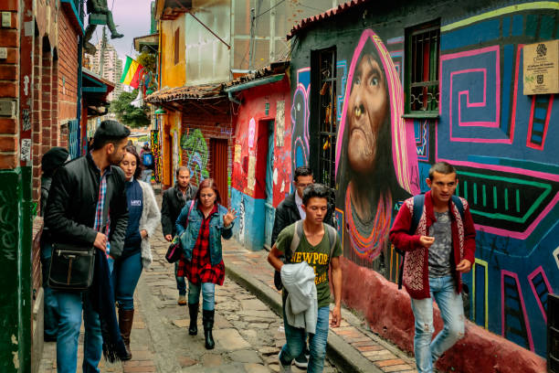 Bogota, Colombia - Tourists and Local Colombians on the Calle del Embudo, in the Historic La Candelaria District of the Andean Capital City Bogota, Colombia - January 27, 2017: Both Tourists and local Colombian people, walk up the narrow Calle del Embudo one of the most colorful streets in the historic La Candelaria district of Bogotá, the Andean capital city of the South American country of Colombia. The street leads to the Chorro de Quevedo, the plaza where it is believed the Spanish Conquistador, Gonzalo Jiménez de Quesada founded the city in 1538. Many walls in this area are painted with either street art, or legends of the pre Colombian era, in the vibrant colours of Latin America. Photo shot on a cloudy morning; horizontal format. calle del embudo stock pictures, royalty-free photos & images