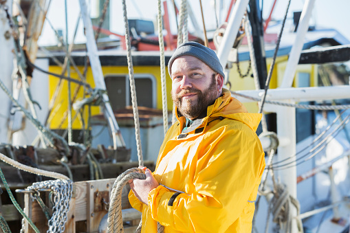 A mature man in his 40s wearing a yellow raincoat, standing on the deck of a commercial fishing boat. He is holding rope in his hand, smiling at the camera.