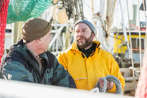 Two mature men in their 40s and 50s standing on the deck of a commercial fishing boat surrounded by nets. The man with the beard and yellow raincoat holding a rope is talking and the other man is smiling.