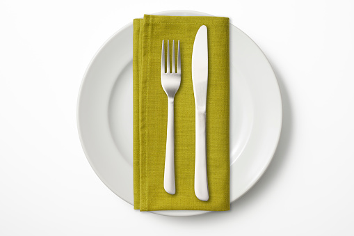 Overhead shot of white plate with green napkin,table knife and fork isolated on white.