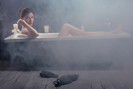 Beautiful woman relaxing in bathtub with lighted candles. Woman taking a bath in steamy bathroom. Looking at camera.