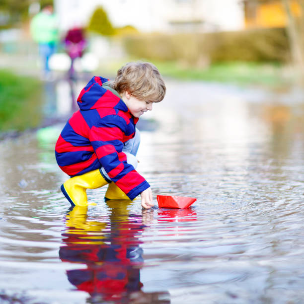 istock Little kid boy playing with paper ship by puddle 652989504