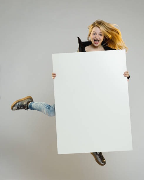 Vertical portrait of excited young woman jumping with a blank poster. Smiling real woman flying with blank billboard over gray background.