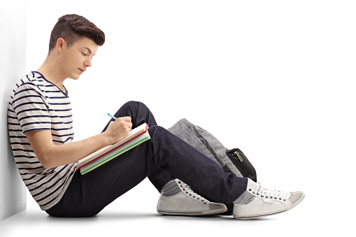 Teen student writing in a notebook and leaning against a wall isolated on white background