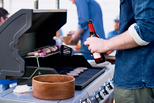 Unrecognizable man with beer bottle standing near grill machine and cooking meat patties