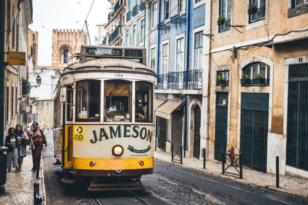 Lisbon, Portugal: Traditional yellow tram moving up on R. Augusto Rosa street. Lisbon cathedral is visible in the background. People are walking on a cobblestone pavement.