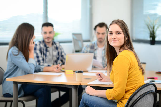 porträt einer jungen frau in freizeitkleidung, die in einem kreativen startup-firmenbüro mit kollegen im hintergrund arbeitet - adult student stock-fotos und bilder