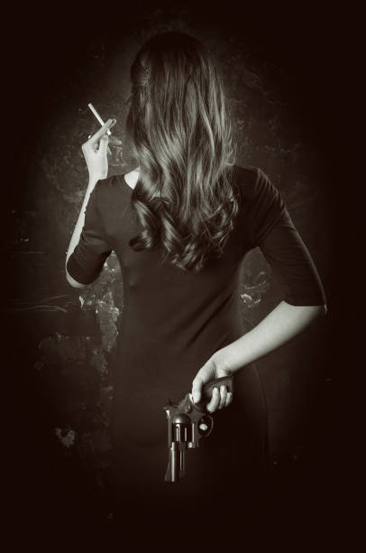 Black and white portrait of dangerous young woman back with long hair, dark dress. She takes gun in her hand Black and white portrait of dangerous young woman back with long hair, dark dress. She takes gun in her hand. femme fatale stock pictures, royalty-free photos & images