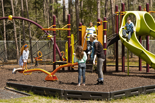 Multi-ethnic group of elementary age school children playing on a school or park playground in spring or summer season.  A teacher or parent is with the children.
