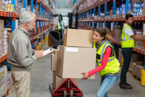 istock Warehouse manager noting on clipboard while female worker carrying cardboard boxes 653217412