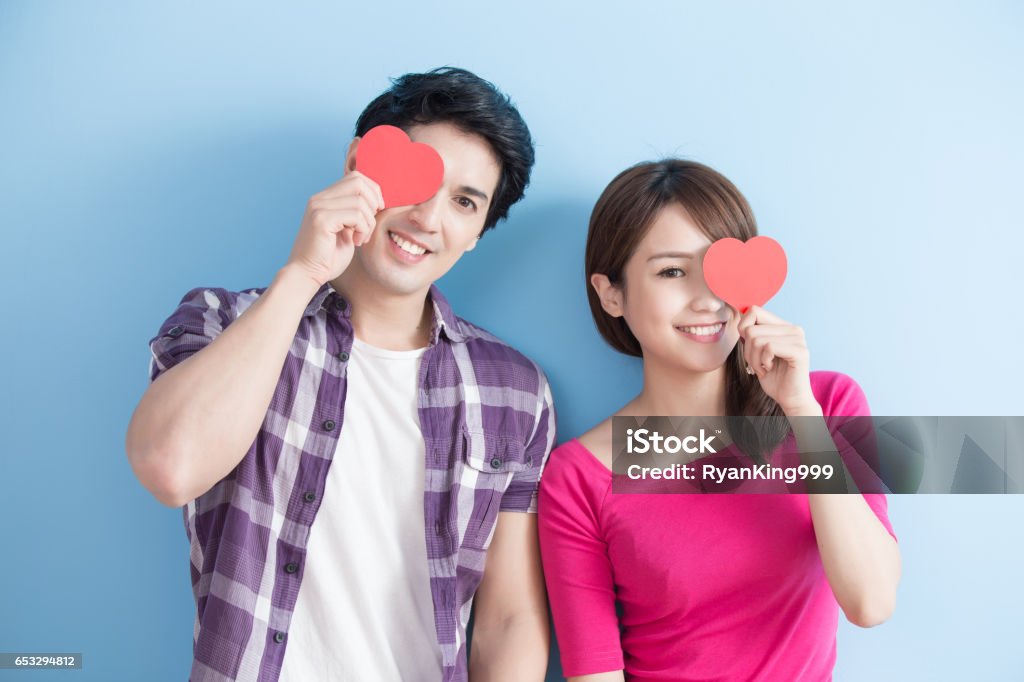Happy couple holding love heart Attractive young couple holding red love hearts over eyes isolated on blue background Chinese Ethnicity Stock Photo