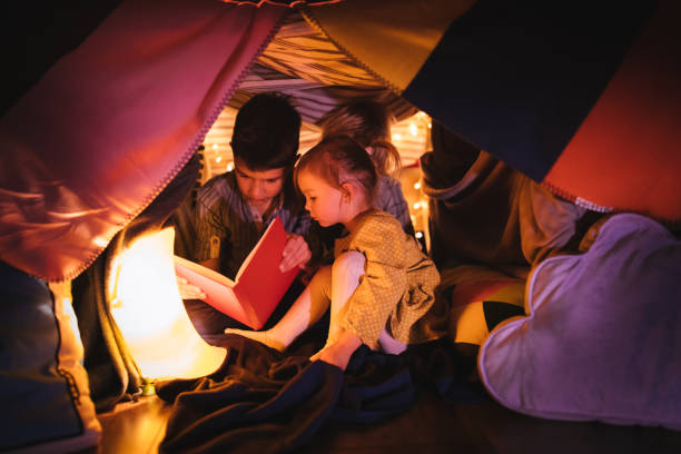 Children reading a story in blanket fort at night One girl and two boys reading a book at night in a blanket tent they built fort stock pictures, royalty-free photos & images
