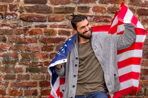 Excited patriot of American style. Excited young man holding American flag while standing against red brick wall during the day, USA Patriotism