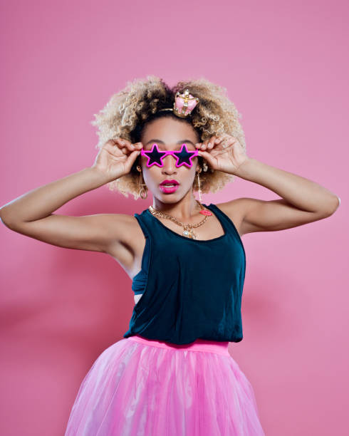 Studio portrait of sensual afro young woman wearing black sleeveless top, pink skirt and star shapd sunglasses. Pink background.
