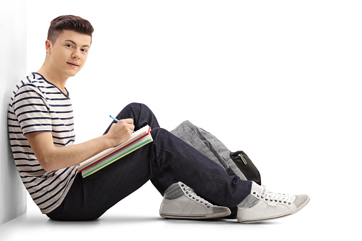 Teenage student leaning against a wall writing in a notebook and looking at the camera isolated on white background