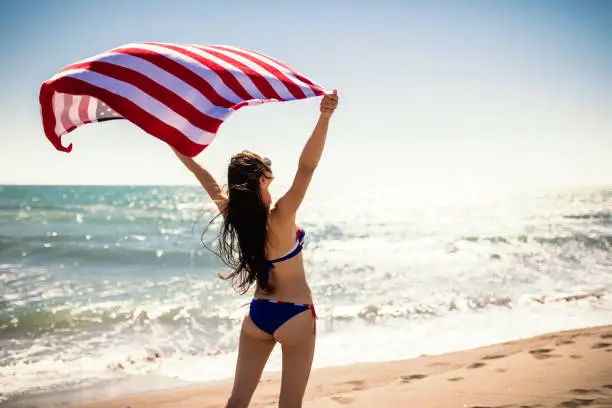 彼女の手でアメリカ国旗とビーチを走るビキニで幸せな若い女 彼女の手でアメリカ国旗とビーチを走るビキニで幸せな若い女