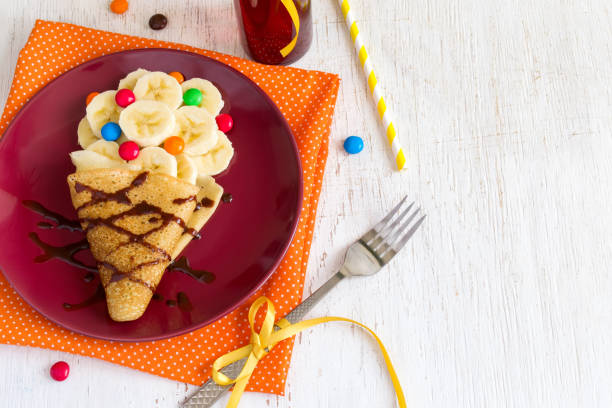 Children's breakfast or dessert - pancake with banana, chocolate topping and colorful candies. Making sweet food in the form of ice cream.