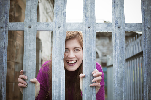 An attractive young caucasian woman laughs as she looks through bars around a ruined building at the derelict docks in Govan, Glasgow, Scotland