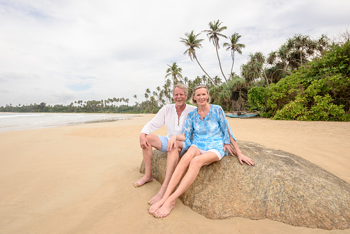 Senior man and woman wearing shorts, on tropical beach with palm trees in background