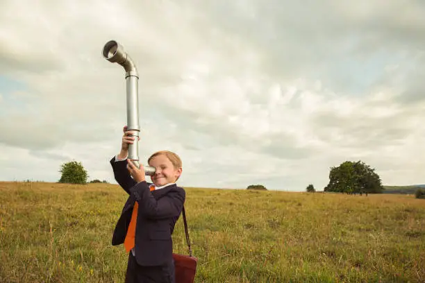 A young British boy dressed in business suit holds a homemade periscope to search for new business opportunities while standing in a rural field in Gloucestershire, United Kingdom. He is searching for new business opportunities.