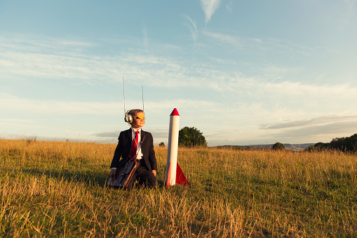 A young child businessman dressed in suit and tie with a unique invention on his head prepares to launch his homemade rocket into the sky.  The boy is counting down the seconds to liftoff in a field in Gloucestershire, England.