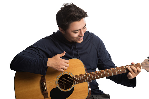 Smiling young man wearing a blue hoodie strumming on a acoustic guitar. Sitting down in front of a white background.