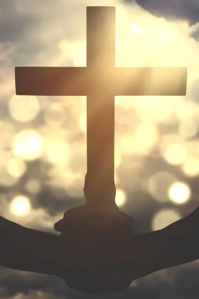 Silhouette of human hands holding a christian crucifix symbol with defocused sunlight on the sky