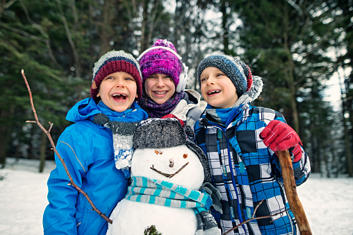 Portrait of little girl and little boys posing with a snowman. Kids are laughing and embracing and having a lot of fun.\n