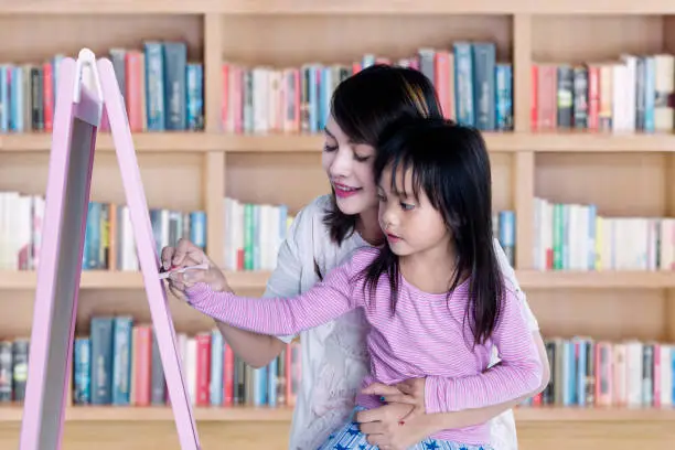 Cute little girl learns to write on blackboard with her teacher, shot in library with bookshelf background