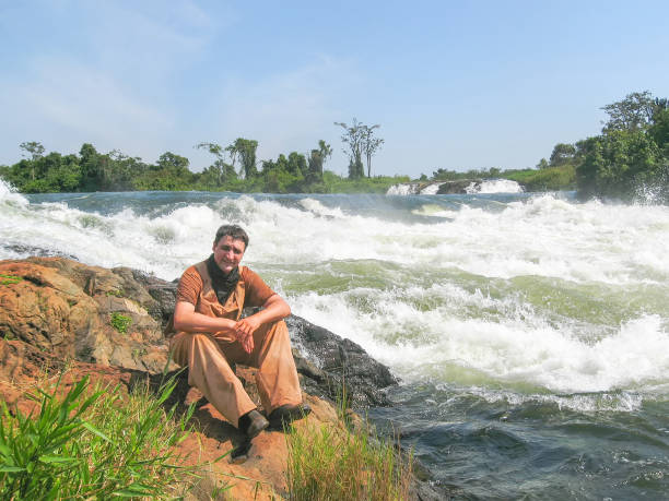 istock Man sits on bank against Victoria Nile River rapids background 653871202