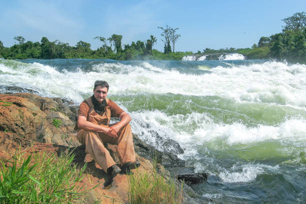 istock Man sits on bank against Victoria Nile River rapids background 653871300