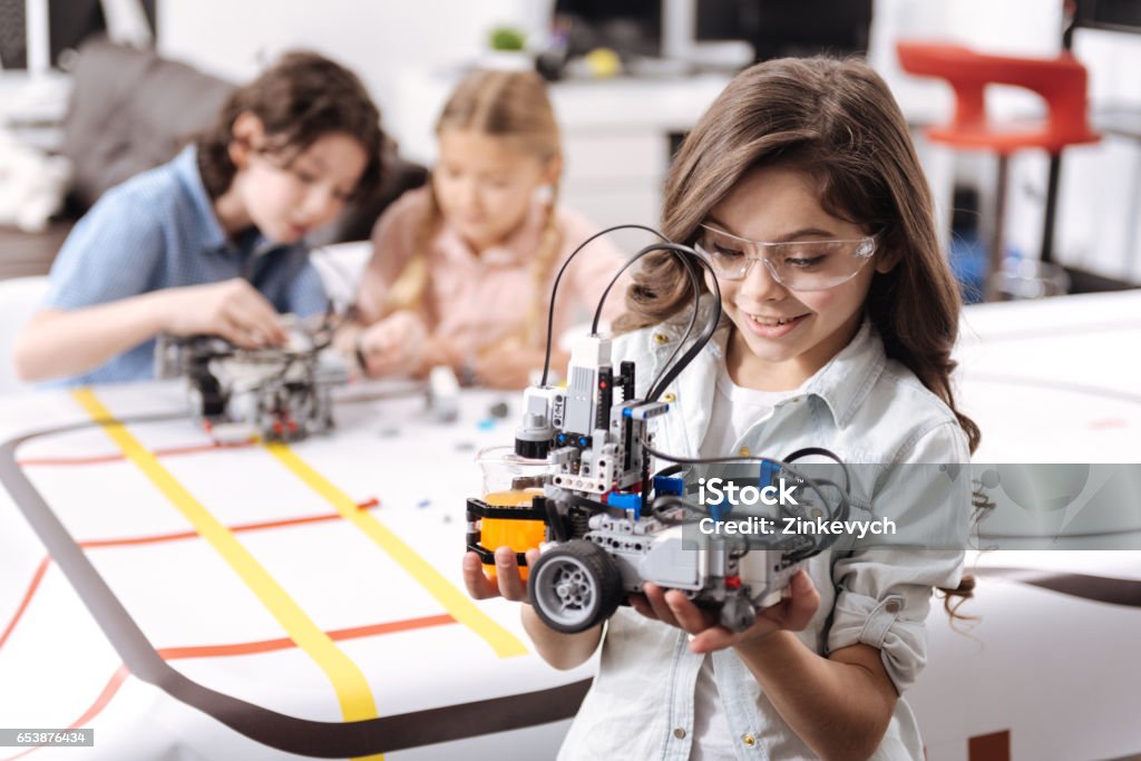 Lovely little girl holding robot at school Smart generation exploring science. Sincere smiling emotional girl standing at school and holding electronic robot while her colleagues working on the project Child Stock Photo