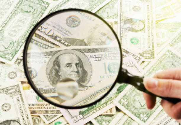 istock Hand holds magnifying glass over $100 bill among mixed dollars 653893642