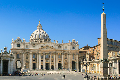 St. Peter's Basilica is an Italian Renaissance church in Vatican City and the papal residence within the city of Rome, Italy. St. Peter's Square Obelisk, sightseeing tourists, street lamps and clear blue sky are in the image.