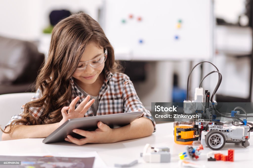 Amused teen girl using tablet in the science studio Getting new knowledge. Smiling involved clever girl sitting in the science studio and using tablet while surfing the Internet Child Stock Photo