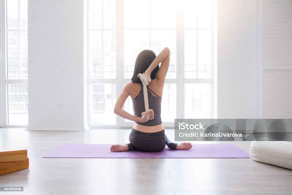 Fitness young woman doing yoga exercise in sportswear Fitness young woman doing yoga exercise in sportswear with strap near a window in studio, back view. Pilates and stretching concept Yoga Stock Photo