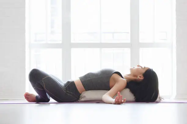 Beautiful young yoga girl lying in asana Shavasana or Corpse Pose using bolster. Fitness woman resting after working out, doing yoga exercise near a window on gym