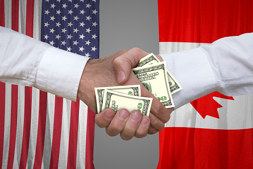 Closeup photo of a 100 dollar bills handshake with USA and Canadian flags background.