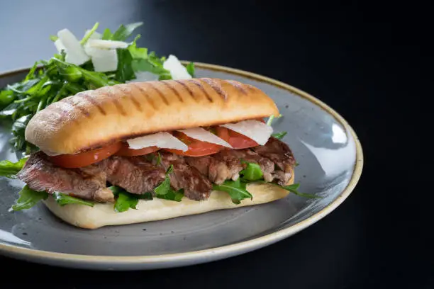 grill marks ciabatta bread sandwich filled with cooked medium beef steak meat tomato salad and parmesan cheese shavings salad on side