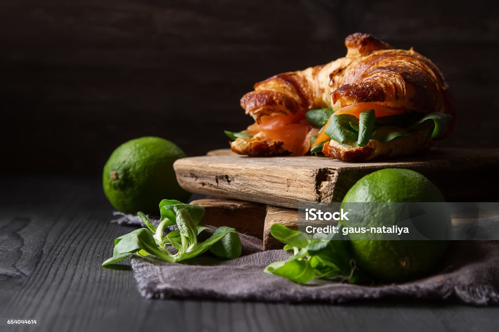 French food for breakfast. Baked Croissant sandwich with salmon French food for breakfast. Baked Croissant sandwich with salmon and fresh salad leaves, green ripe lime on dark wooden board. Dark wood background Close-up Stock Photo