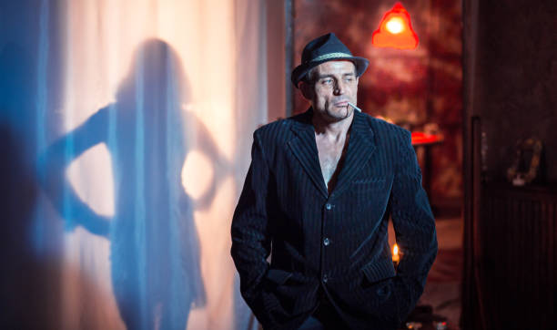 Charismatic mature man dressed in striped gangster jacket on his bare torso. The man in a hat with a cigarette in his mouth is standing with his hands in his pants pockets. He pensive looking away. Silhouette of slim woman in short dress behind a white curtain is a backgroung for the man. Full-length mirror in which the room is reflected behind the man. Studio shooting in retro style