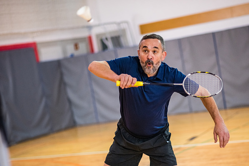 Mature man hitting shuttlecock with badminton racquet in court.