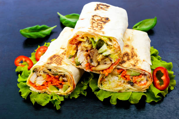 istock Shawarma - Middle Eastern dish made from lavash (pita), stuffed with chicken, mushrooms, fresh vegetable salad, sauce. Serving on lettuce leaves on a black background. 654112914