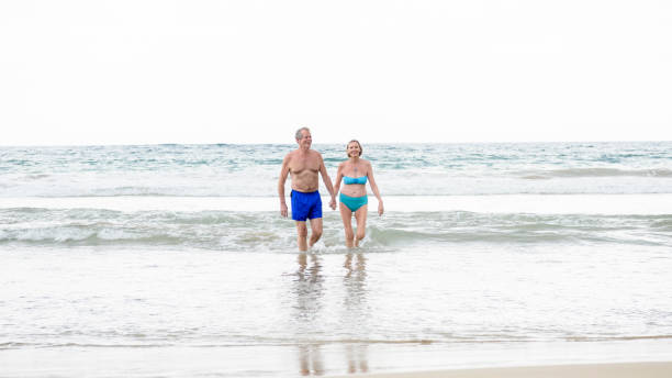 vista frontal de la pareja senior en mar usar traje de baño, las manos - swimming trunks bikini swimwear red fotografías e imágenes de stock